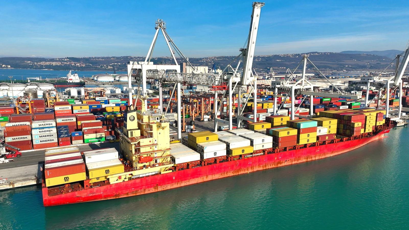 Red container ship loaded with colourful containers berthed under cranes at Port Koper
