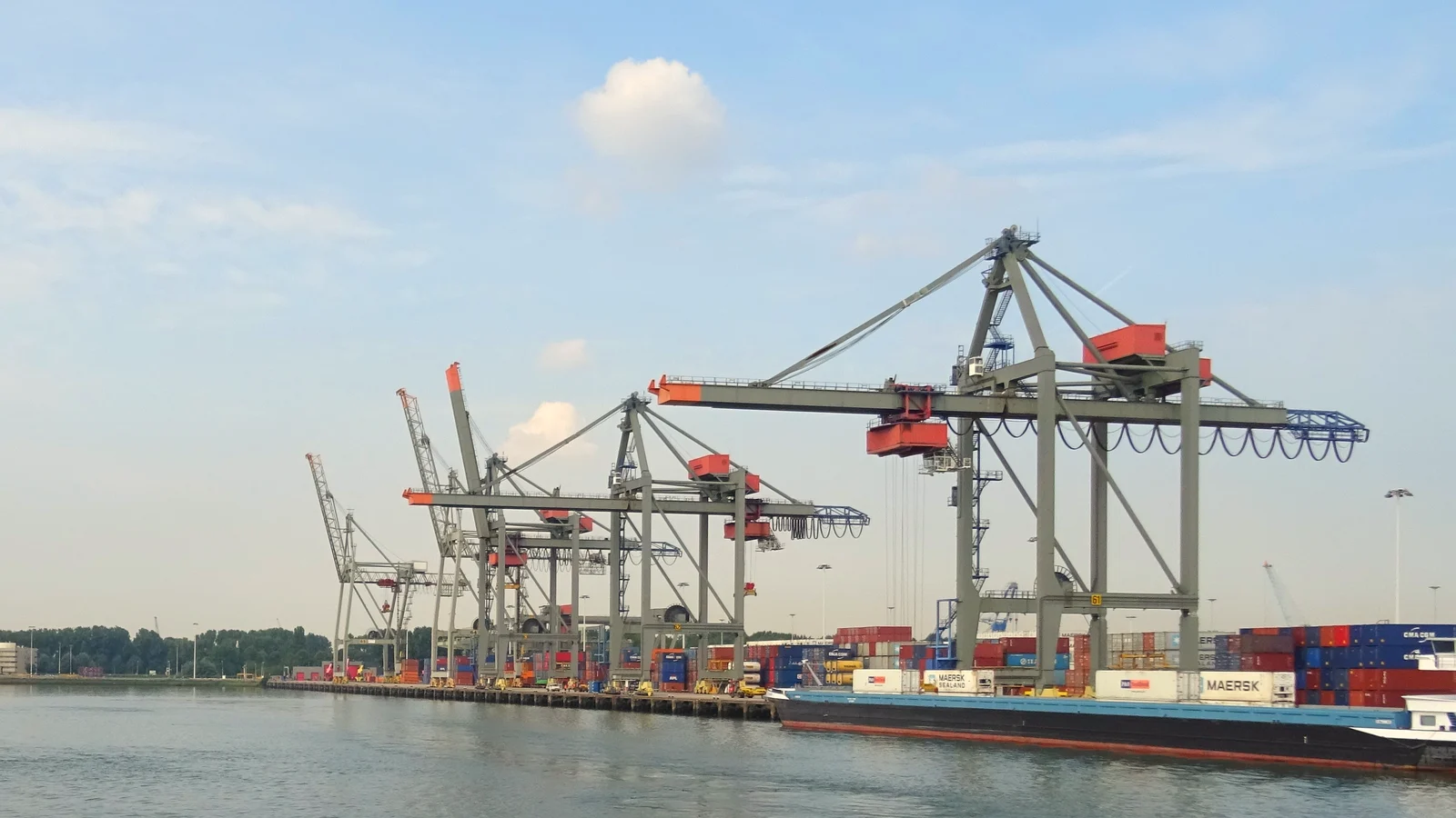 Ship-to-shore container cranes and a loaded barge at a Rotterdam terminal under blue sky