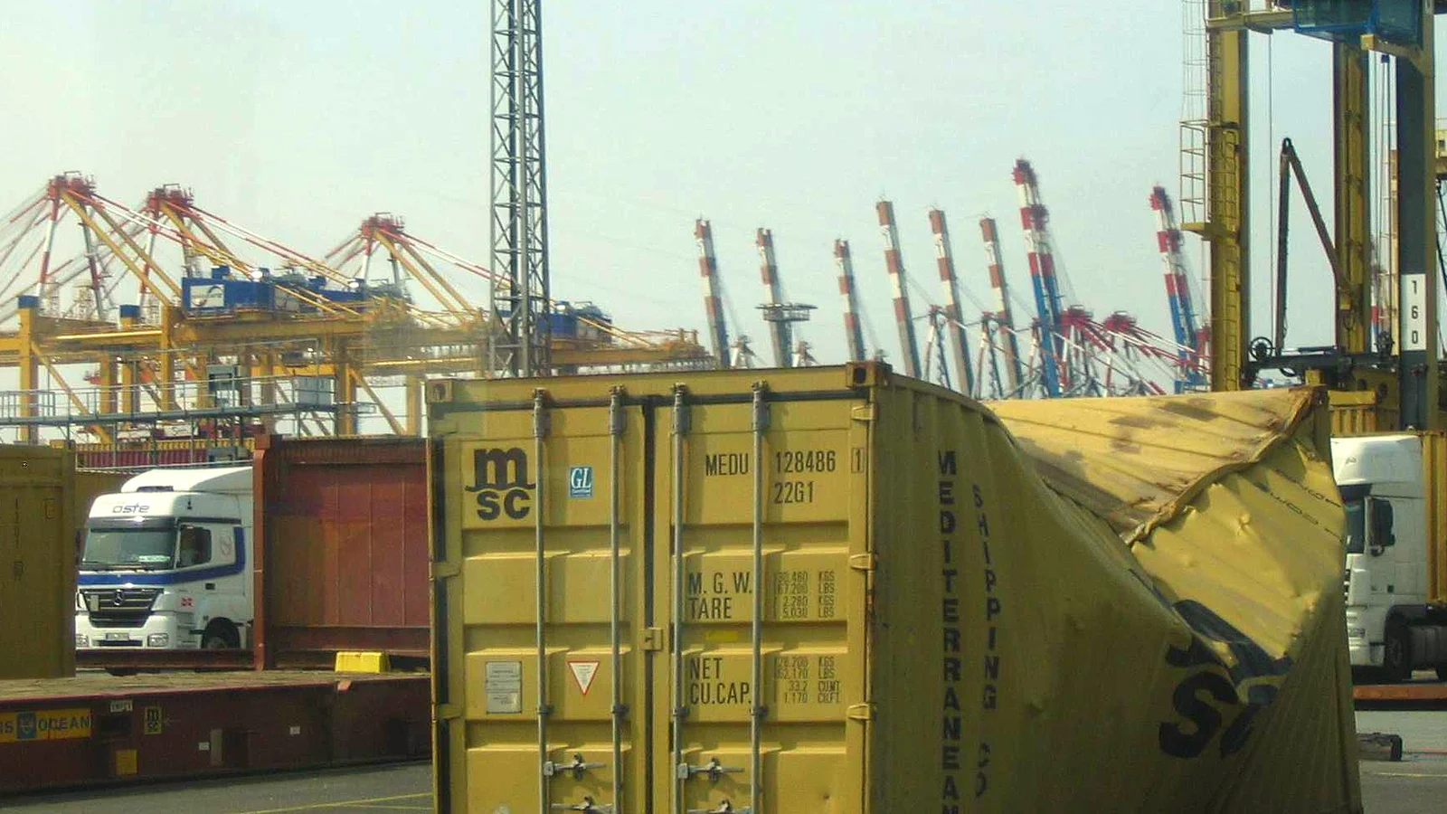 Damaged MSC shipping container with a crushed side at a port terminal, cranes behind