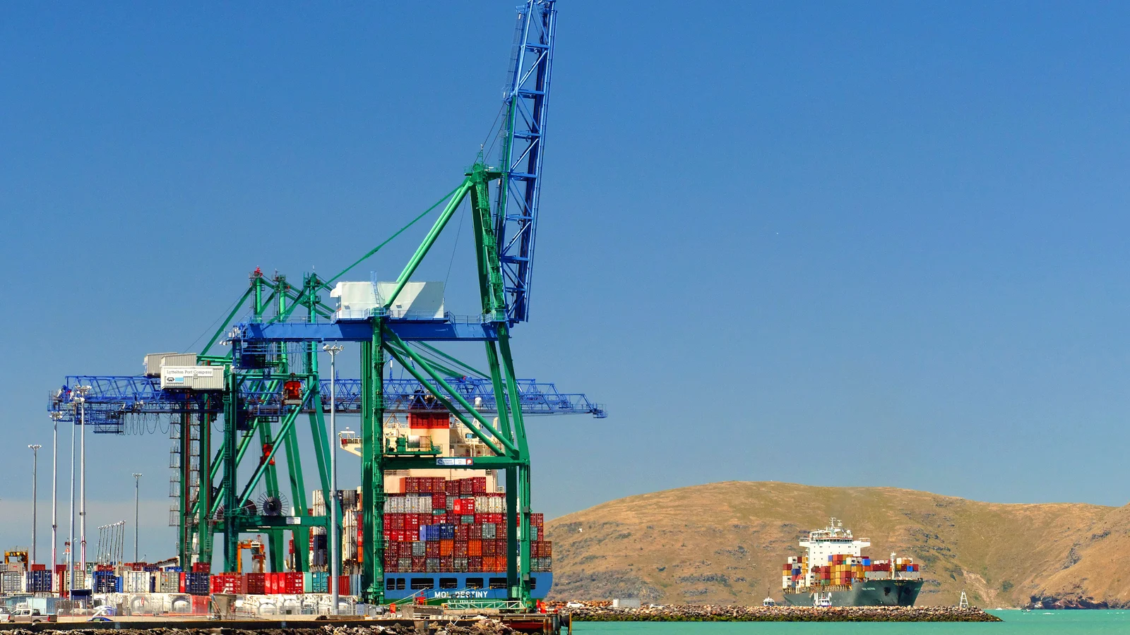 Green ship-to-shore crane over container stacks at Lyttelton port, ship and hills behind